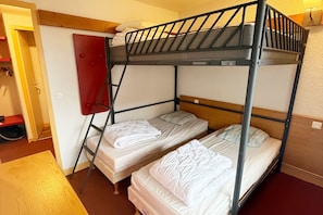 Habitació amb un llit búnquer, dos llits, una escala, una porta vermella i una cadira de fusta.