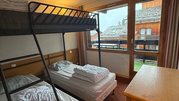 A room with bunk beds, a wooden desk, and a window with a view of buildings.
