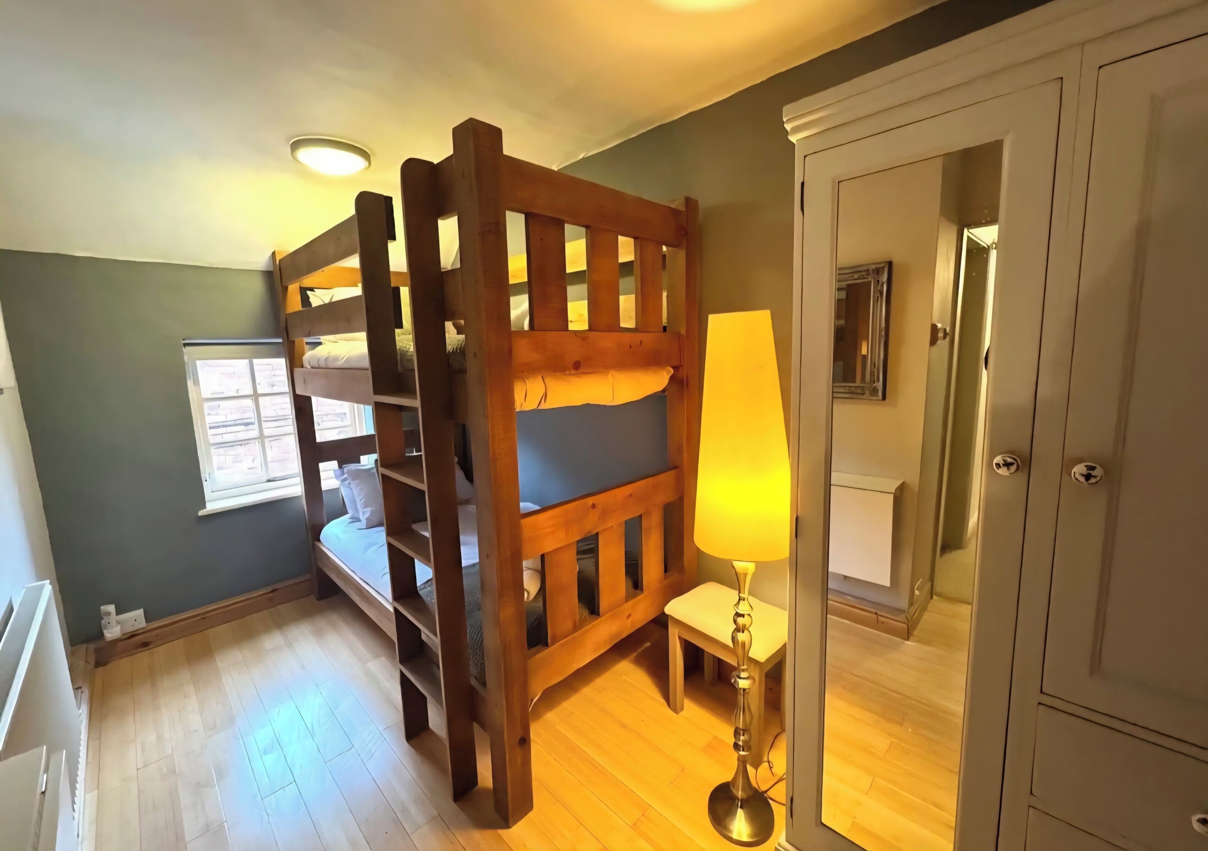 Un lit superposé avec une structure en bois, une table de chevet avec une lampe et une armoire dans une pièce dotée d’un plancher en bois et d’une fenêtre.