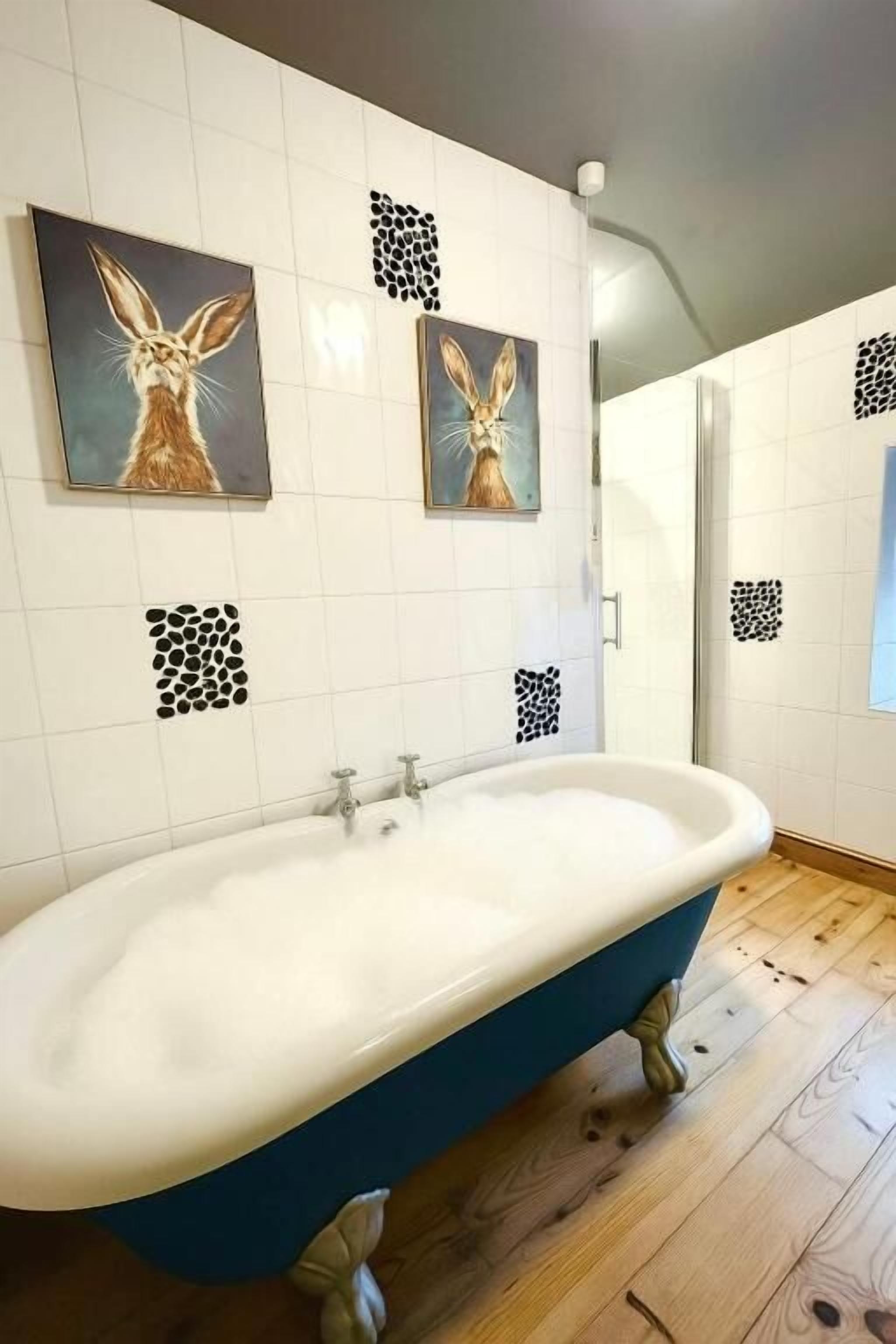 Une salle de bain avec une baignoire sur pieds, des murs recouverts de carreaux blancs et deux tableaux sur le thème des lapins.