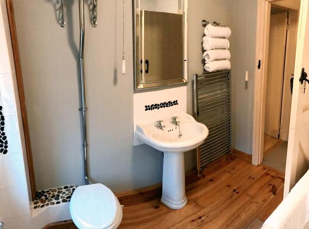 Une salle de bain avec un lavabo sur colonne blanc, une douche et des toilettes. On y trouve un porte-serviettes avec des serviettes pliées et un miroir au-dessus du lavabo.