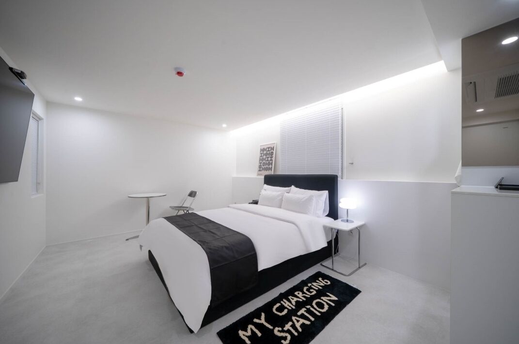 A modern bedroom with a bed, bedside table, and a charging station sign on the floor.