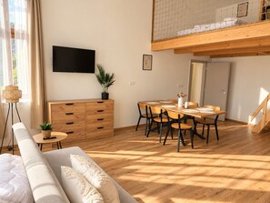 A modern living room with a sofa, a dining table with chairs, a TV mounted on the wall, and a staircase leading to an upper level.