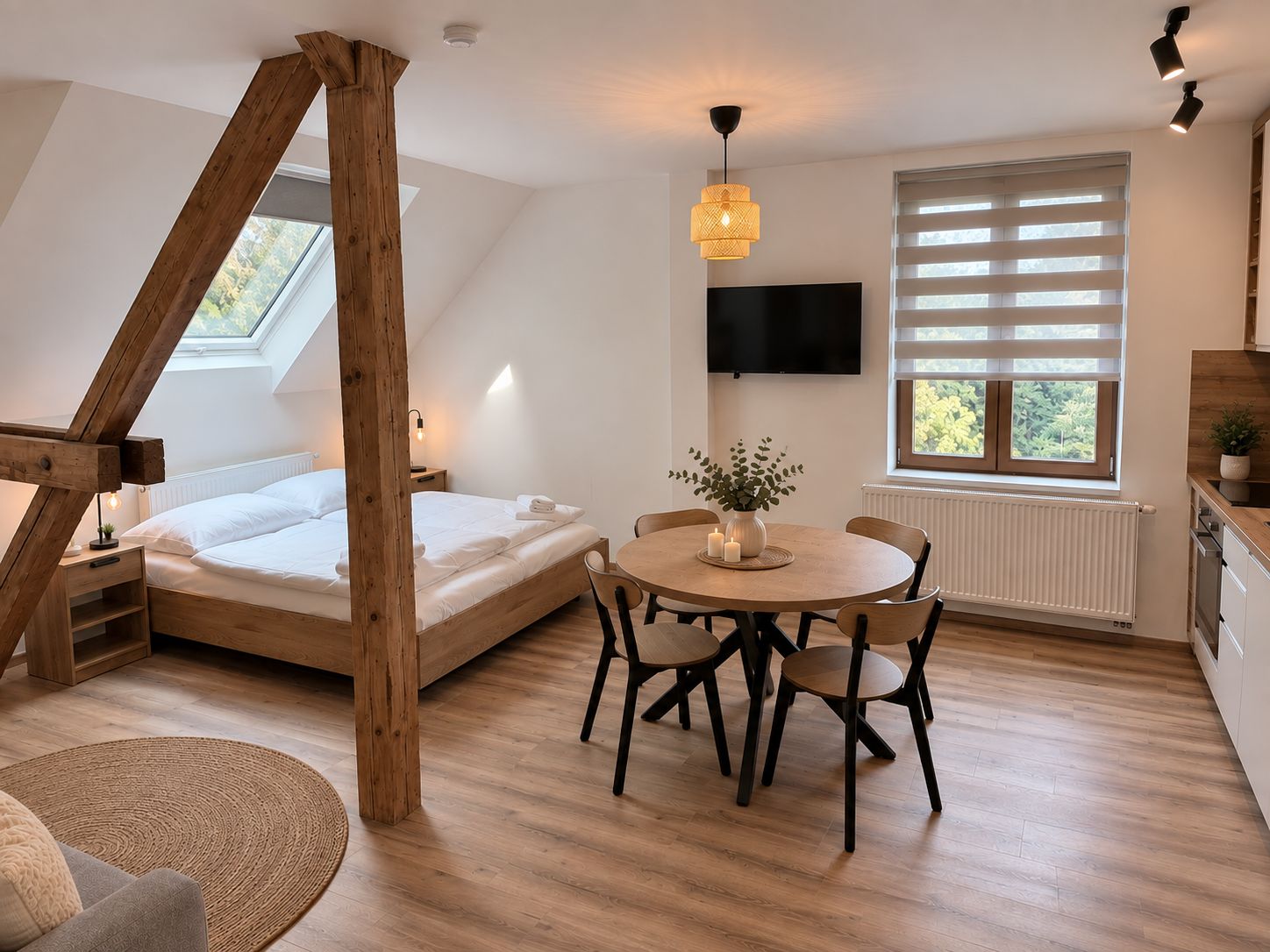 A bedroom with a sloped ceiling, a bed, a round dining table with chairs, a television mounted on the wall, and a window with blinds.
