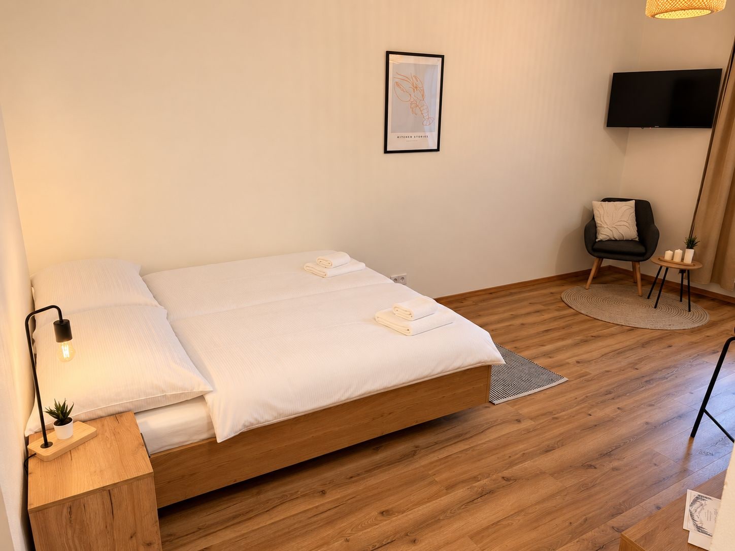 A minimalist bedroom with a wooden bed, a bedside lamp, a framed picture on the wall, a chair, and a small table with candles.