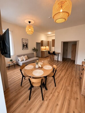 A modern living room with a round wooden dining table, a sofa, a TV mounted on the wall, and a kitchen area in the background.