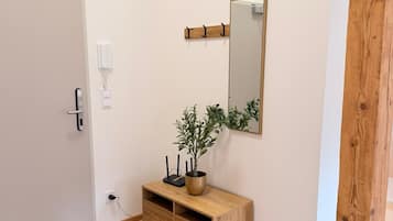 A hallway with a wooden floor, a white door, a small wooden console table with a plant, a wall-mounted mirror, and a coat rack.