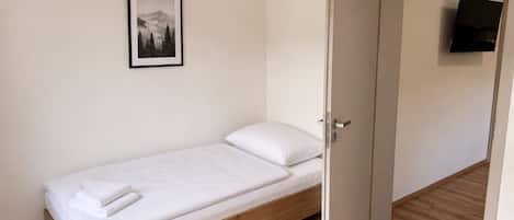 A single bed with a wooden frame and white bedding, a door leading to another room, and a framed picture on the wall.