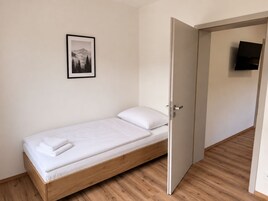 Una cama individual con estructura de madera y ropa de cama blanca, una puerta que da a otra habitación y un cuadro en la pared.