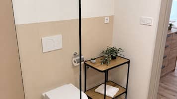 Un baño con inodoro, un pequeño estante de madera con plantas y una zona de ducha.