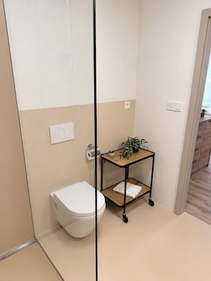 A bathroom with a toilet, a small wooden shelf with plants, and a shower area.