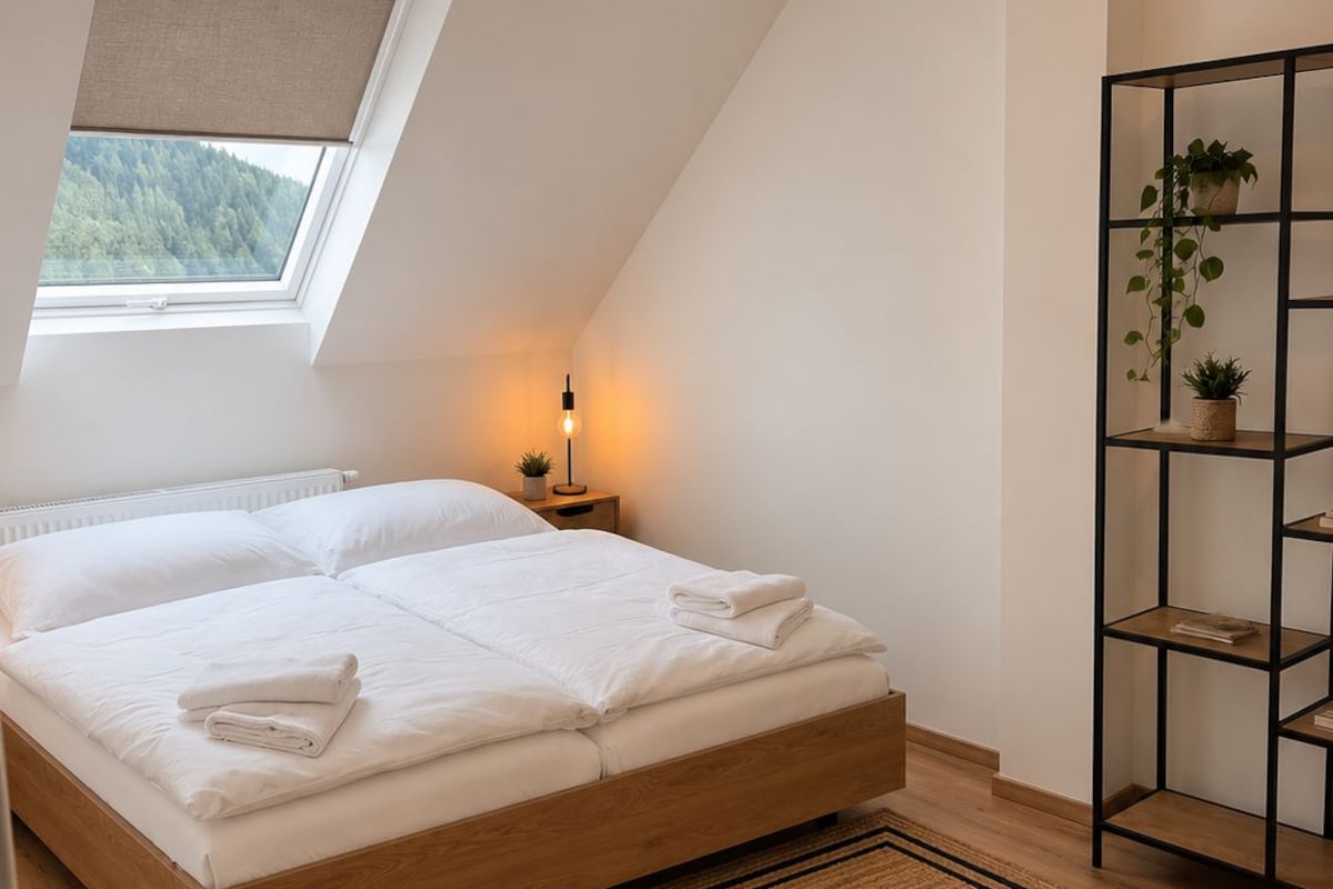 Un dormitorio con techo inclinado, tragaluz, cama con ropa blanca, mesita de noche con lámpara, piso de madera y estante con plantas.