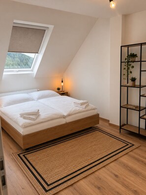 A bedroom with a sloped ceiling, a skylight, a bed with white linens, a bedside table with a lamp, a wooden floor, and a plant shelf.