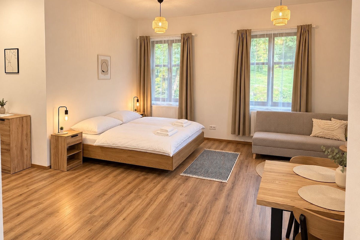 A bedroom with a wooden floor, a bed with white linens, a sofa, a desk with a lamp, and two windows with curtains.