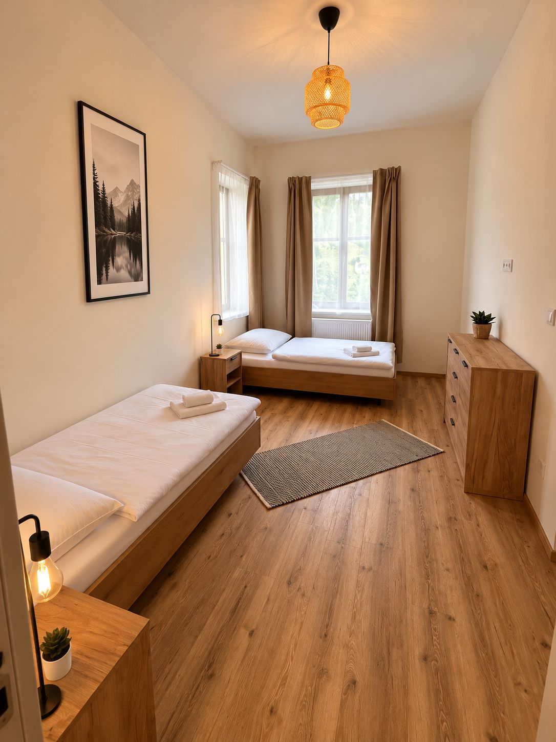 Et minimalistisk soveværelse med to senge, en trækommode, en hængende lampe og et billede i ramme på væggen.