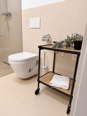 A modern bathroom with a toilet, a shower, a wall-mounted toilet paper holder, a small shelf with a towel, and a potted plant.