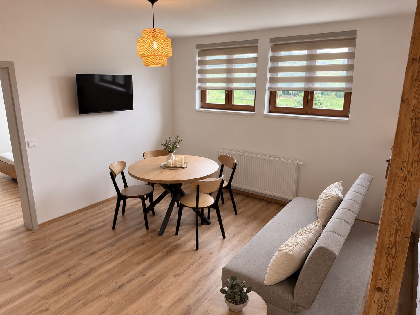Un espacio de estar moderno con una mesa de comedor redonda, cuatro sillas, un sofá con cojines, un televisor montado en la pared y dos ventanas con persianas.