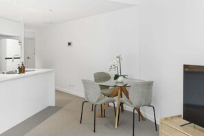 A modern kitchen with a white countertop, a built-in oven, and a sink. There is a dining area with a glass table and four grey chairs. A flat-screen TV is mounted on the wall to the right. - NEW! City Centre One-Bedroom Apartment; Sleeps 4 (Brisbane)