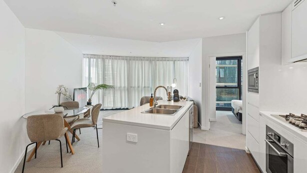 A modern kitchen with white cabinetry, a central island with a sink, and a dining area with chairs and a table. - NEW! City Centre One-Bedroom Apartment; Sleeps 4 (Brisbane)