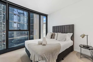 A modern bedroom with a large bed, a headboard, a bedside table, and a window with a city view. - NEW! City Centre One-Bedroom Apartment; Sleeps 4 (Brisbane)