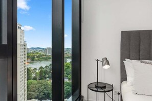 A hotel room with a large window, a bedside table with a lamp, and a bed with white bedding. - NEW! City Centre One-Bedroom Apartment; Sleeps 4 (Brisbane)
