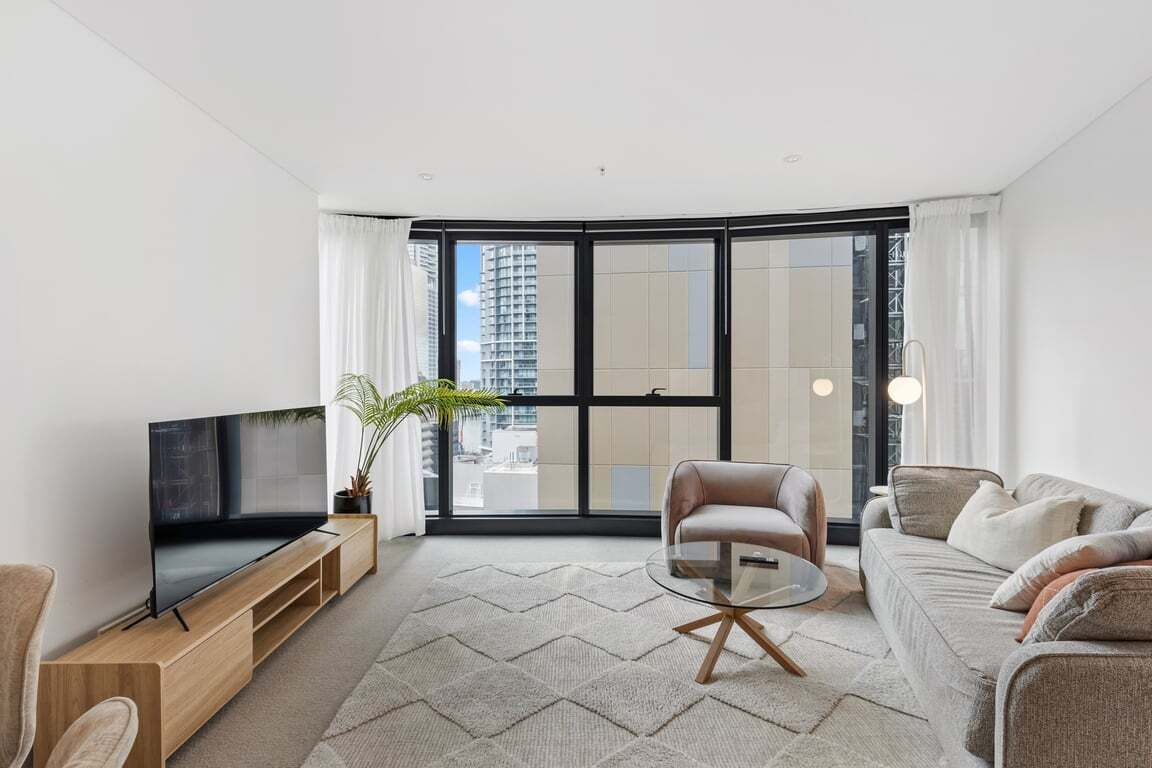 A modern living room with a flat-screen TV, a glass coffee table, a sofa, and a large window offering a city view.
