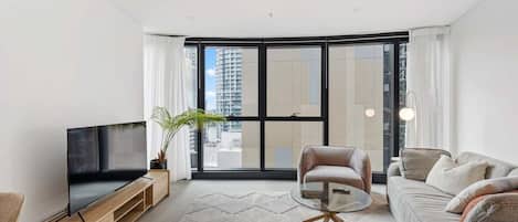A modern living room with a flat-screen TV, a glass coffee table, a sofa, and a large window offering a city view.