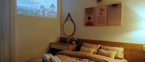 Hotelkamer met een bed, nachtkastje met een lamp, spiegel aan de muur en een projectiescherm waarop een afbeelding van een band te zien is.