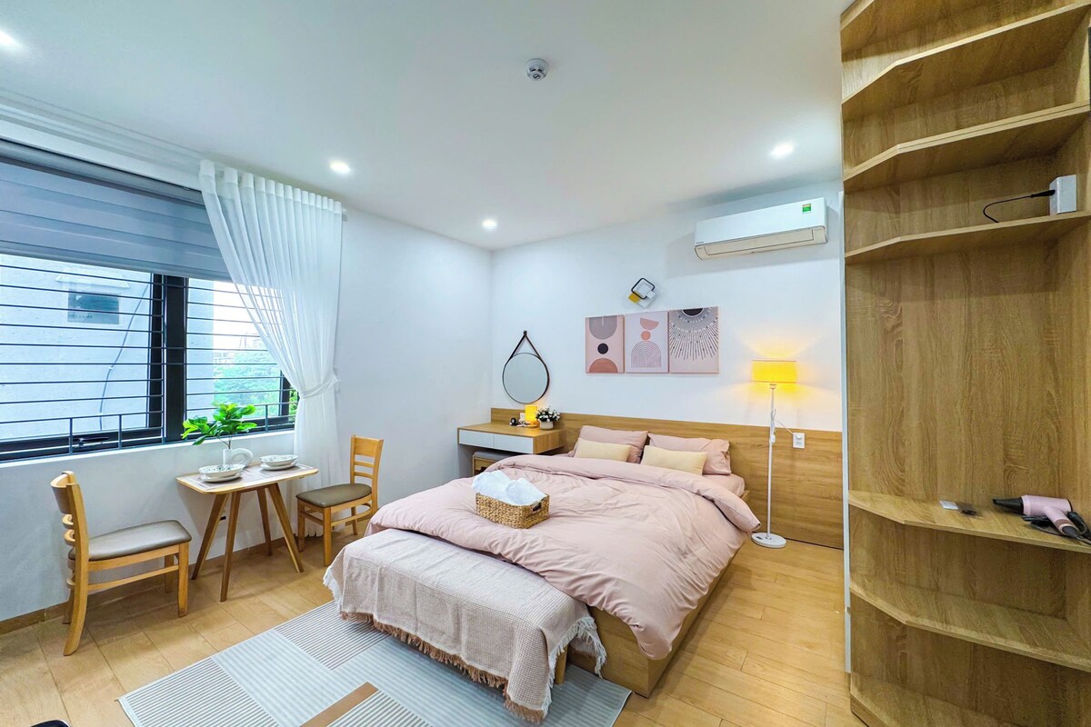 A bedroom with a bed, a desk, a chair, a bedside table, a lamp, and an air conditioning unit.