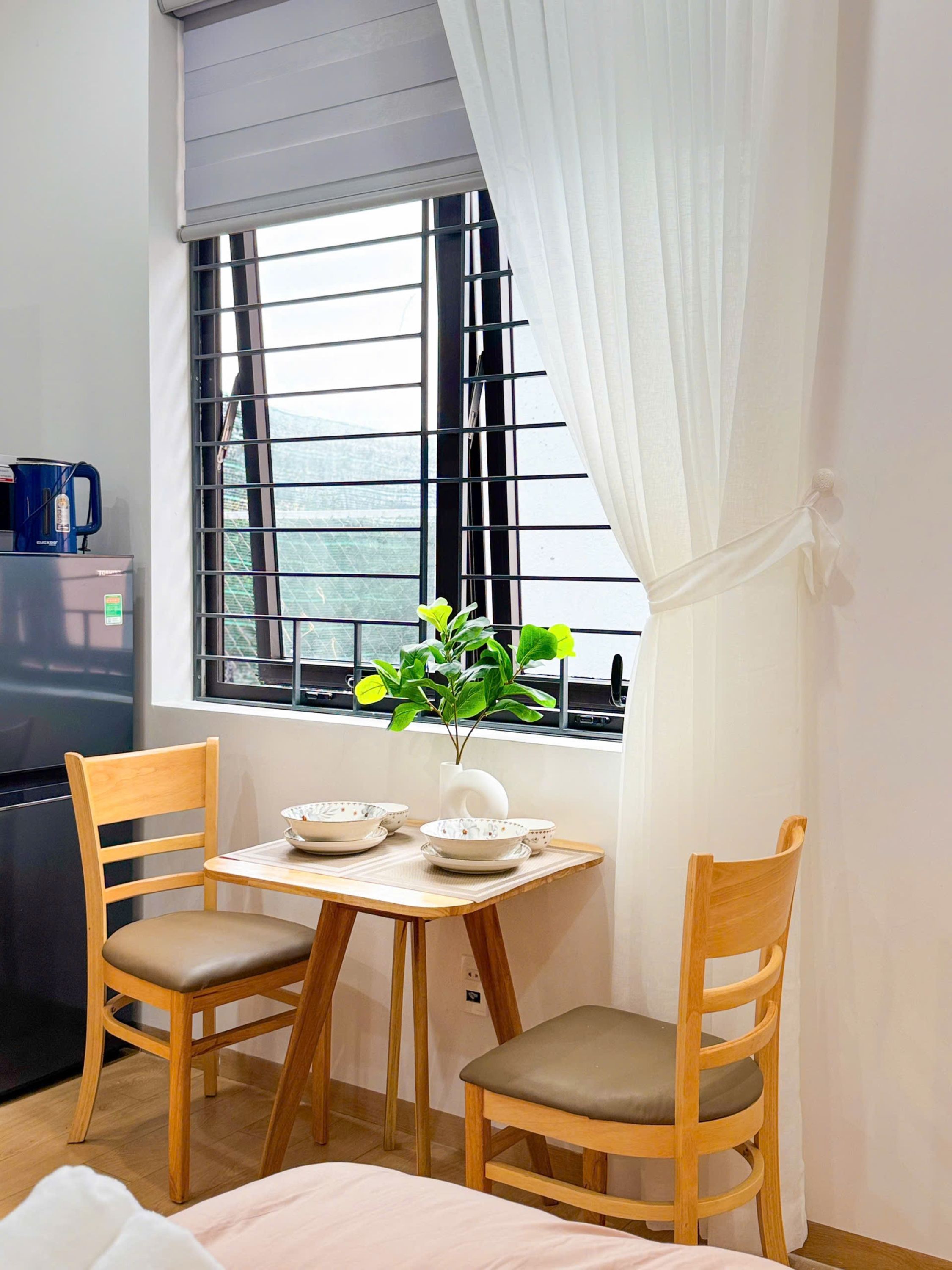 A small dining area with a table, two chairs, a potted plant, and a window with metal bars.