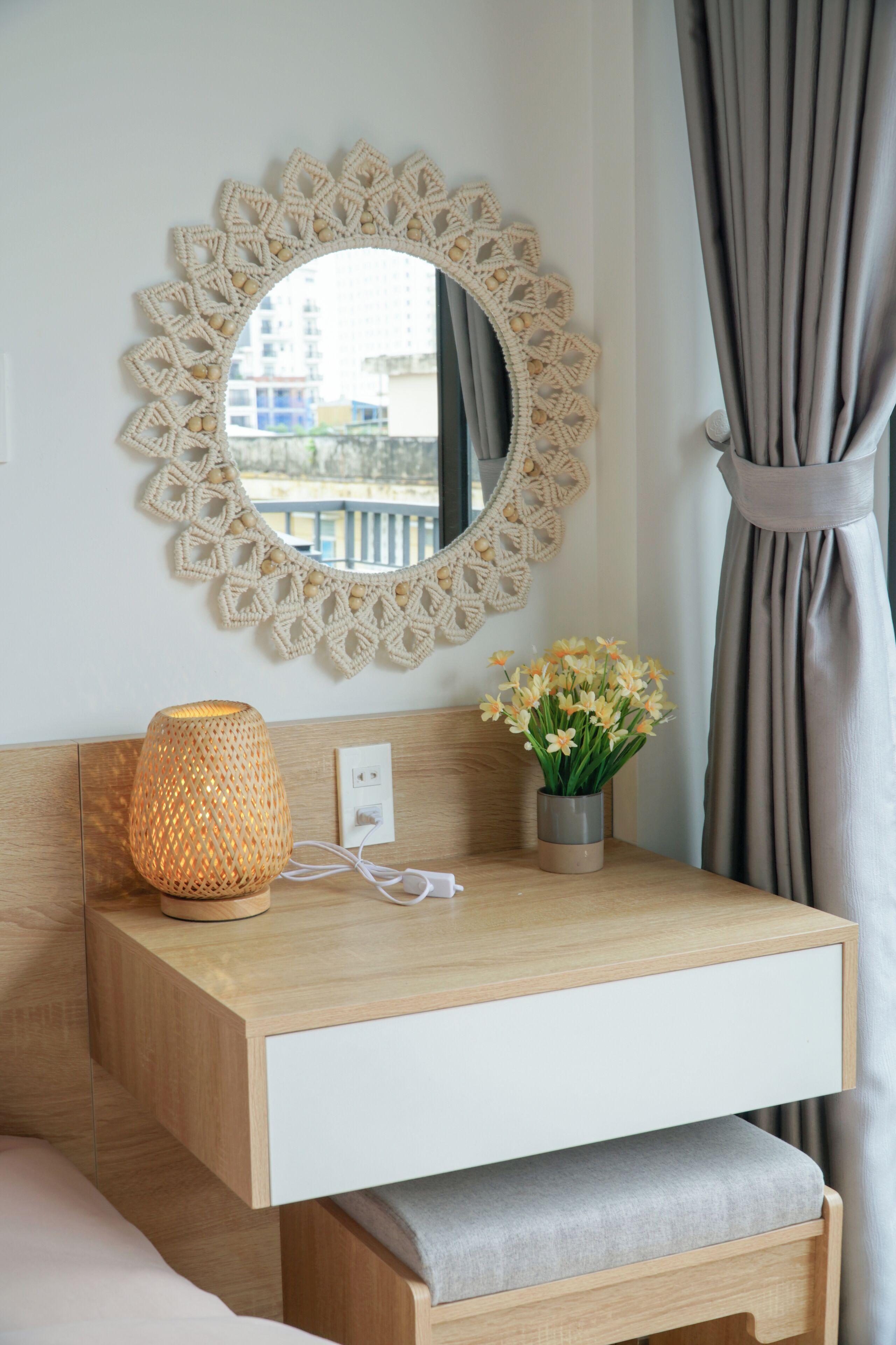 A hotel room with a wooden bedside table, a woven lamp, a vase with flowers, a round mirror, and a view of buildings through the window.
