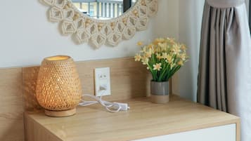 A hotel room with a wooden bedside table, a woven lamp, a vase with flowers, a round mirror, and a view of buildings through the window.