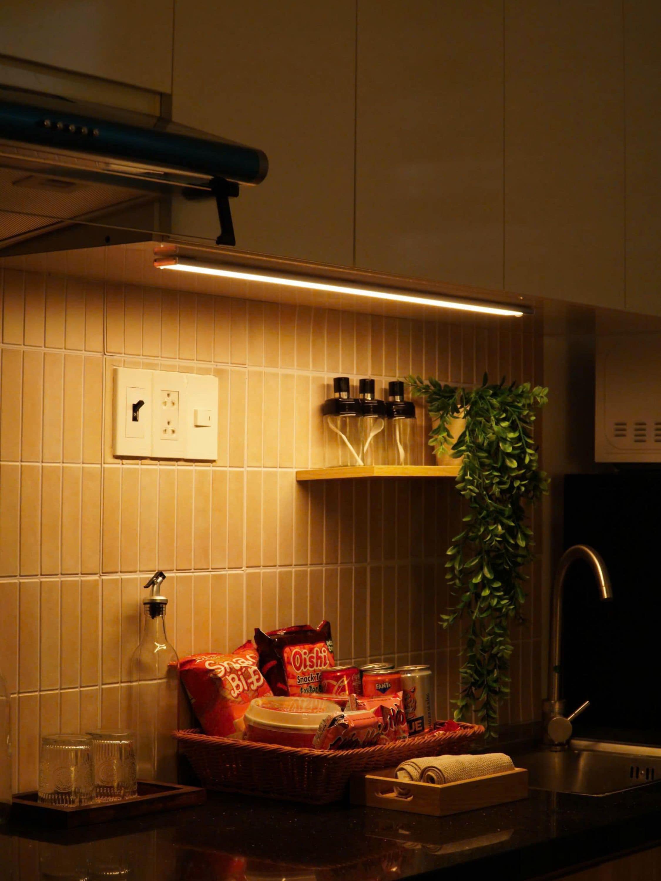 A kitchen with a tiled backsplash, a shelf with bottles, a potted plant, and various food items.