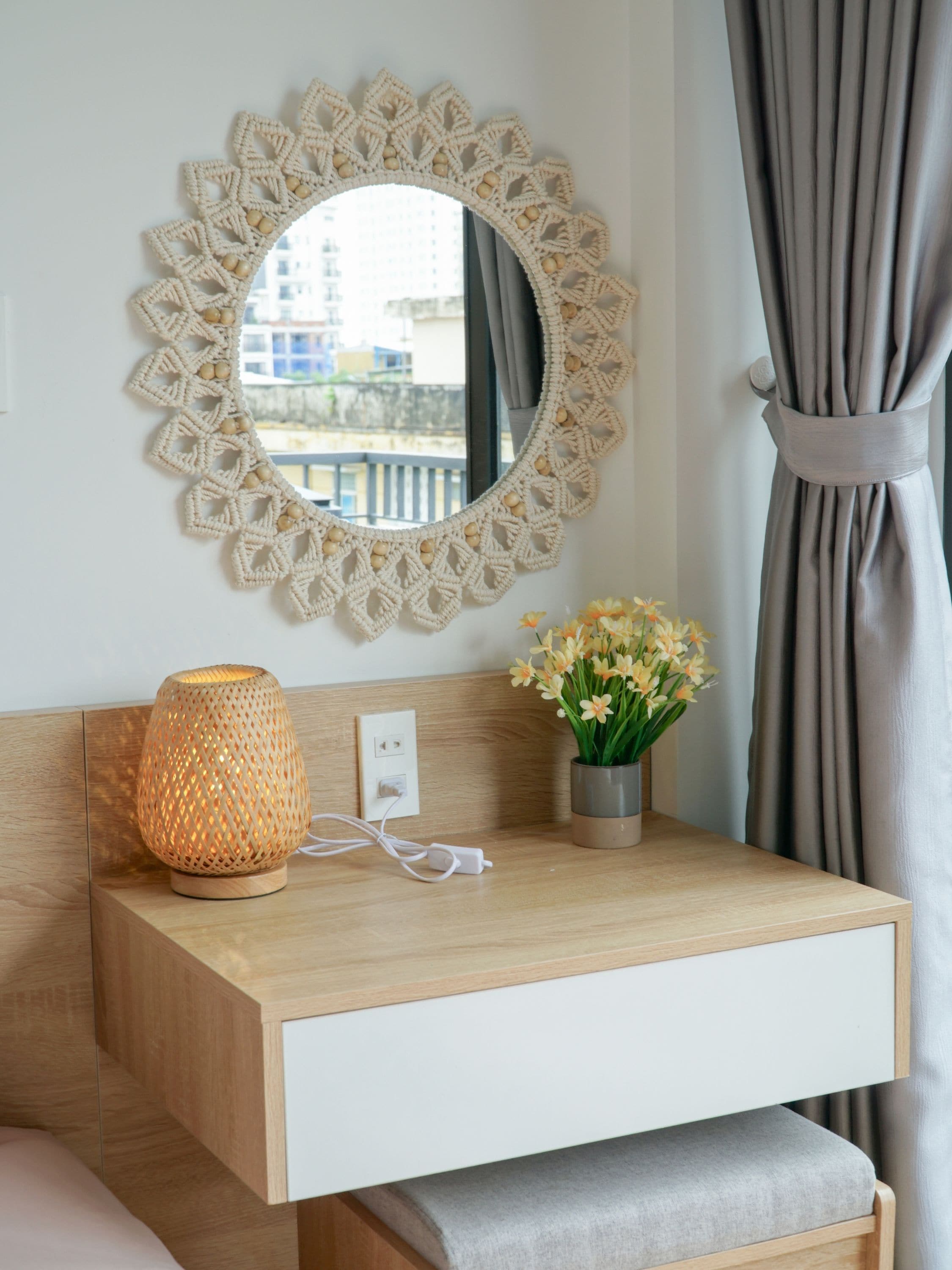 Una mesita de noche de madera con un espejo, un macramé colgado en la pared, una lámpara y un florero con flores.