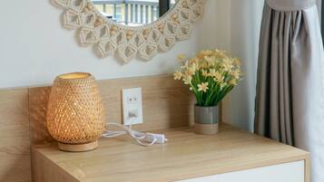 Una mesita de noche de madera con un espejo, un macramé colgado en la pared, una lámpara y un florero con flores.