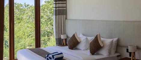 A hotel room with a large bed, a bedside table with a lamp, and a view of greenery through the window.