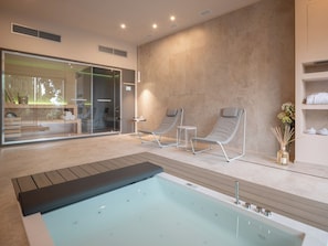 Un área de piscina interior moderna con pared de vidrio, sillas de descanso y un jacuzzi.