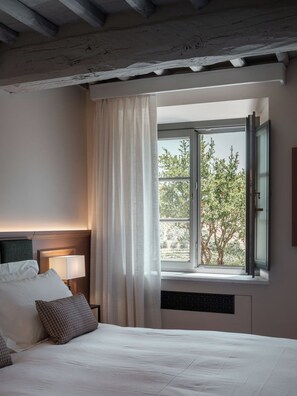 Un dormitorio con cama, mesita de noche, lámpara, ventana con cortinas y vistas a árboles.