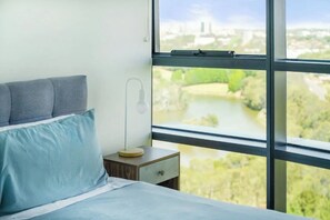 A bedroom with a bed, a nightstand, a lamp, and a window with a view of a river and greenery. - Park View 2BR With Secure Parking | Near to Mall (Sydney Olympic Park)
