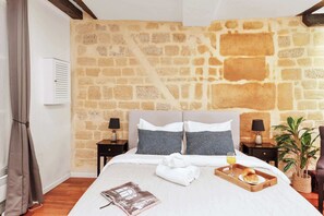 A hotel room with a stone wall, a bed with white bedding, two bedside tables with lamps, a potted plant, and a tray with breakfast items. - Cozy Studio in the Heart of Paris - Louvre (Paris)