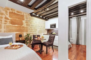 Ein Zimmer mit Steinwand, einem Bett mit Frühstücksbrettchen, einem Tisch mit Stühlen und einer Küchenzeile mit Mikrowelle.