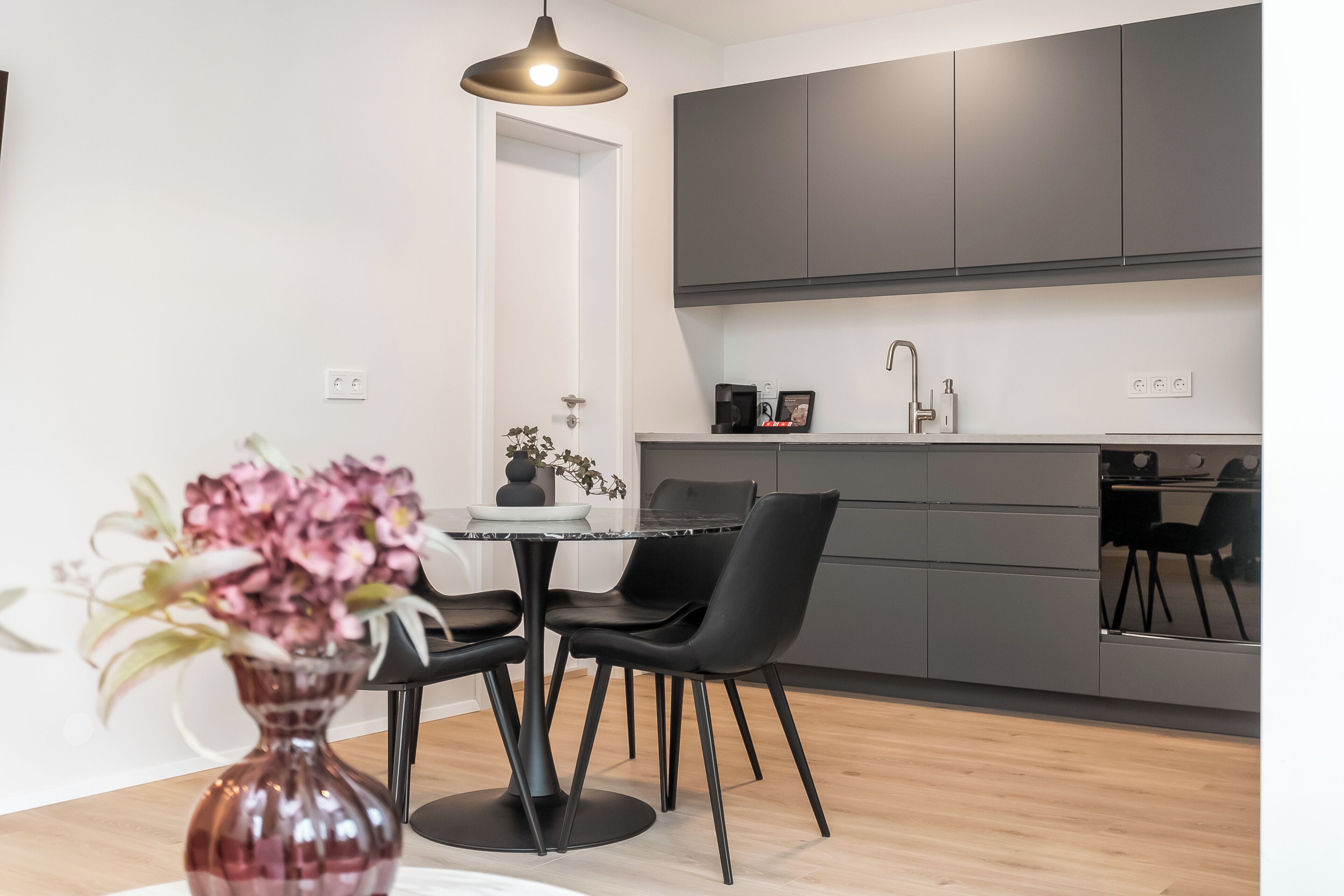 Una cocina moderna con armarios oscuros, un comedor con mesa y sillas negras, y un florero con flores.