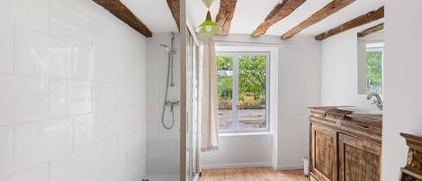 A bathroom with white tiled walls, a wooden floor, a shower with a handheld showerhead, a window with a view of greenery, and a wooden vanity with a mirror.