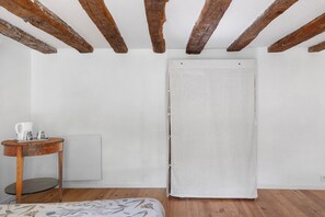A room with exposed wooden beams, a white towel hanging on a door, a small round table with a white pitcher and cups, and a radiator on the wall.