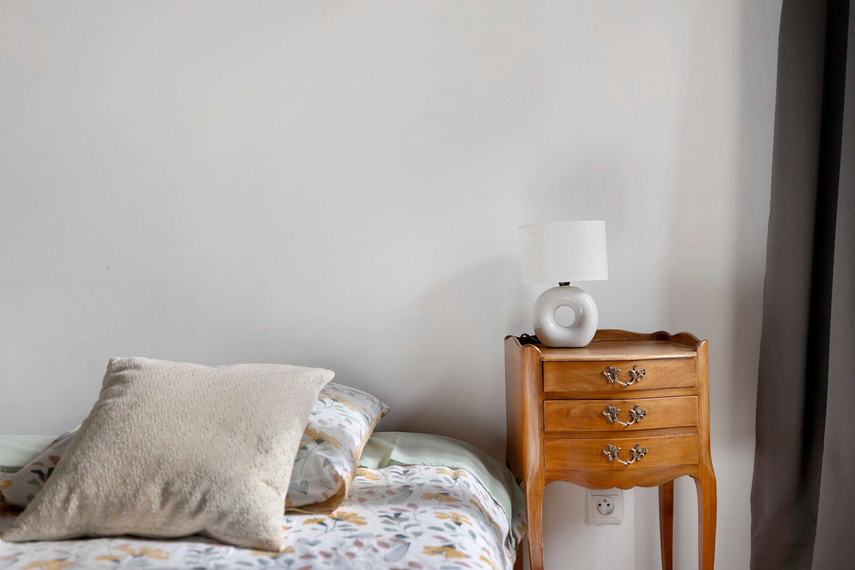 A neatly made bed with a floral-patterned bedspread, a single white pillow, and a bedside table with a white lamp and a wooden drawer.