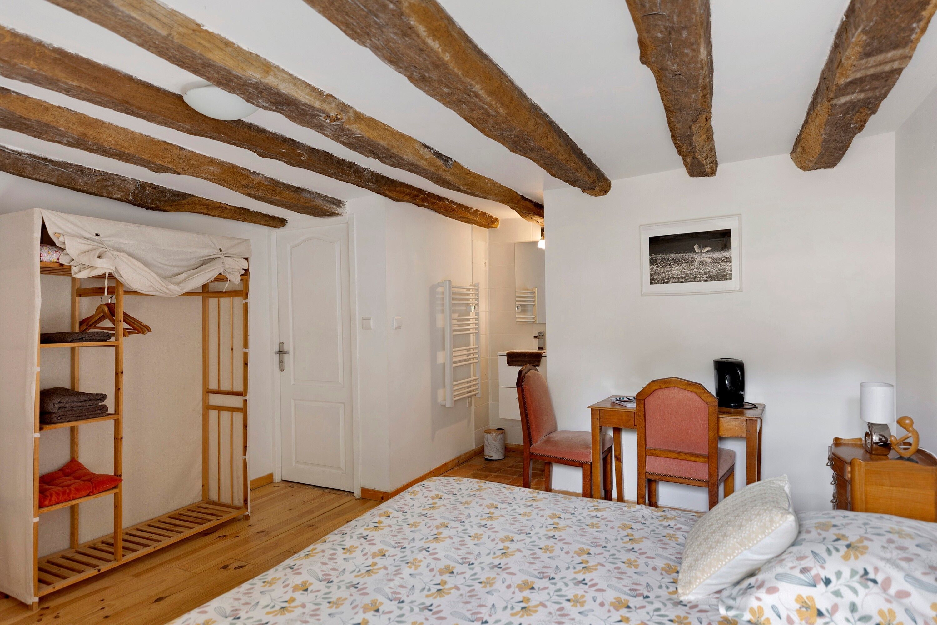 A bedroom with a wooden ceiling, a bed with a floral bedspread, a chair, a table with a kettle, and a radiator.