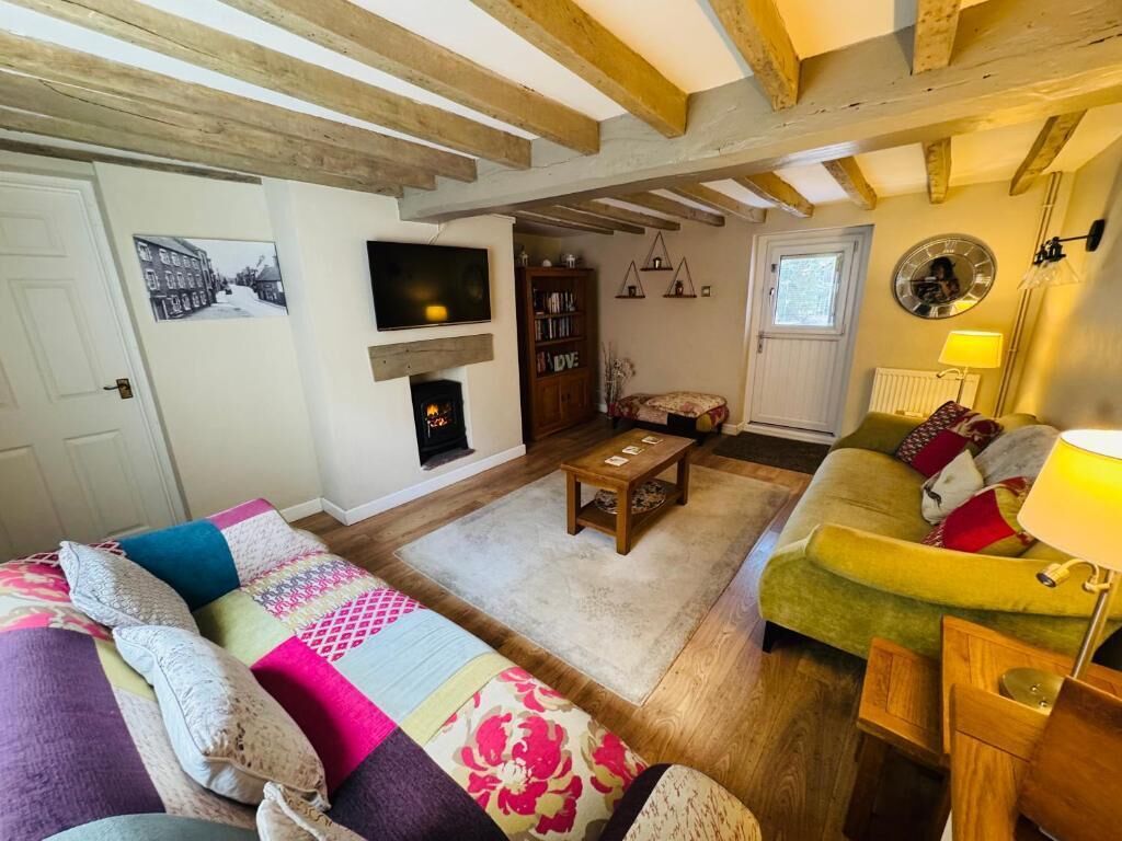 A cozy living room with a fireplace, wooden beams, a sofa, a green armchair, and a bookshelf.
