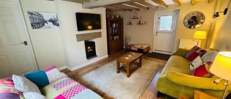 Una sala de estar acogedora con chimenea, vigas de madera, un sofá, un sillón verde y una estantería.