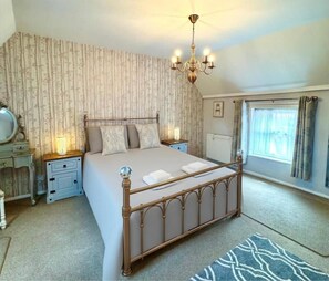 Un dormitorio con cama con dosel, candelabro, mesita de noche con lámpara, ventana con cortinas y alfombra estampada.
