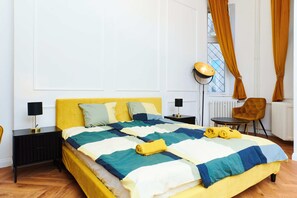 A bed with a yellow headboard, a bedside table with a lamp, a chair, and a window with curtains. - Refined Central Sanctuary – Perfect Romantic Getaway by the River – 701 (Budapest)